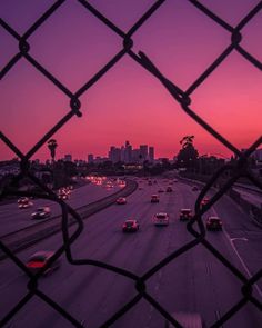 cars driving down the highway at sunset through a chain link fence with city buildings in the background