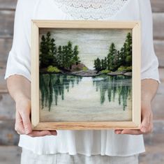 a woman holding a painting in her hands with water and trees painted on the frame