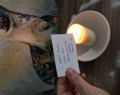 a person holding a piece of paper next to a candle on a table with a painting