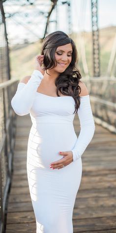 a pregnant woman poses on a bridge while wearing a white dress and heels for her belly