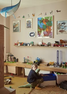 a young boy sitting on the floor in front of a desk and shelves with toys