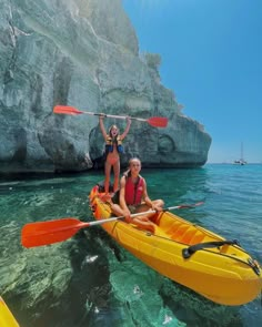 two women in kayaks on the water with rocks behind them and one holding up her paddle