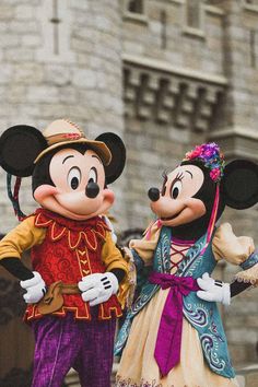 two mickey and minnie mouse characters standing in front of a castle with people dressed up