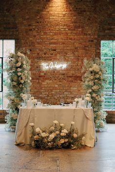 a table with white flowers and greenery on it in front of a brick wall