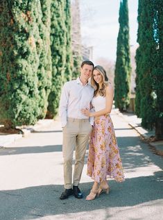 a man and woman standing in front of trees on the sidewalk with their arms around each other