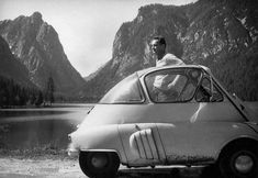 a man sitting in the driver's seat of a small car next to a mountain lake