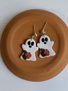 two white ghost shaped earrings with black eyes and red flowers on a brown plate against a white background