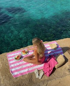 a woman sitting on top of a beach towel next to the ocean with food in front of her