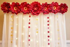 red paper flowers hanging from the side of a white curtain with lights around them and on top of sheer curtains