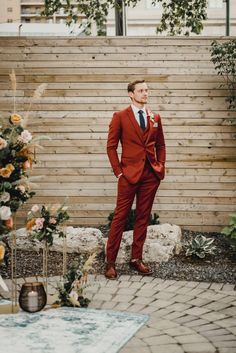 a man in a red suit and tie standing next to a wooden fence with flowers