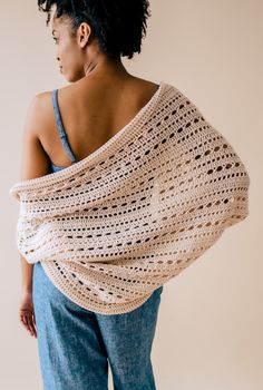 a woman wearing a white crochet shawl and blue jeans is standing in front of a wall