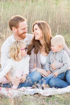 a man and woman with two children sitting on a blanket in the grass smiling at each other