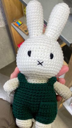 a crocheted white rabbit wearing a green shirt and shorts is held up by someone's hand