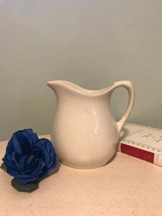 a white pitcher sitting next to a blue flower on top of a table with books