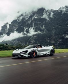 a silver sports car driving down the road in front of a mountain range with clouds