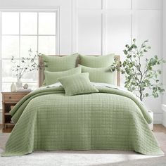 a bed with green comforters and pillows in a room next to a window filled with potted plants