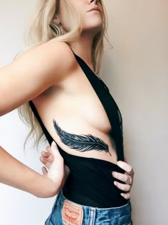 a woman with a feather tattoo on her chest is posing for the camera while wearing jeans and a black tank top