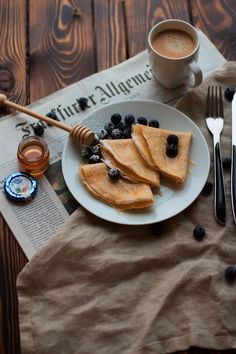 pancakes with blueberries and honey sit on a plate next to a cup of coffee