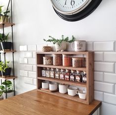 a clock mounted to the side of a white brick wall above a wooden shelf filled with spices