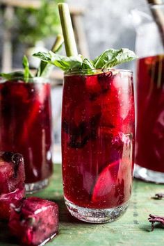 two glasses filled with red liquid sitting on top of a wooden table next to ice cubes