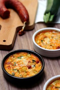 two bowls of food on a table with sausages and other foods in the background