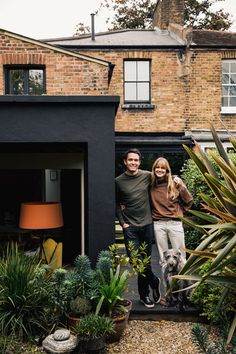 a man and woman standing next to each other in front of a house with plants