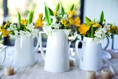 three white vases with yellow and white flowers in them on a tablecloth covered table