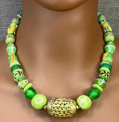 This 20" necklace feels like the green grass of an African summer. It is designed with various handmade Ghana glass beads in shades of green with a large 'basket weave' brass focal bead from Côte d'Ivoire. It has a large brass lobster cloth glass with extension chain for ease of use and positioning on the neck. These beads are all handmade in Africa by crafts people who have been creating these beads for centuries. This creates a sustainable business for many African families using a completely Traditional Green Beaded Necklace For Festival, Green Spiritual Necklace For Festival, Green Spiritual Necklace For Festivals, Green Polished Beads Necklace For Festivals, Green Polished Beaded Necklace For Festival, Traditional Green Beads For Festival, Traditional Green Adjustable Necklace, Unique Green Necklace For Festivals, Unique Green Necklace For Festival