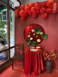 a red table topped with a vase filled with flowers next to a mirror covered in paper lanterns