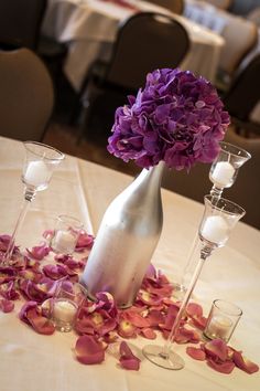 purple flowers in a silver vase surrounded by wine glasses and candles on a white table cloth