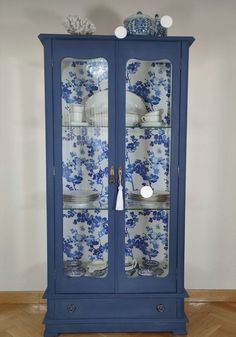 a blue china cabinet with glass doors and flowers on the front, sitting in a wood floored room