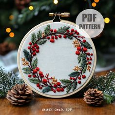 an embroidered christmas wreath with red berries and pine cones is displayed on a wooden table