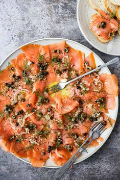 a white plate topped with lots of salmon and olives next to bread on top of a table