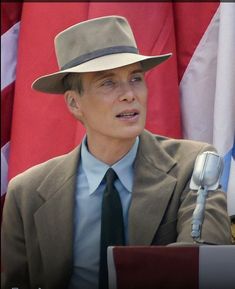 a man in a suit and tie sitting at a podium with flags behind him wearing a hat