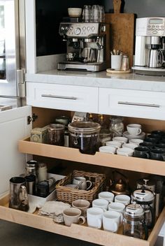 an open drawer in a kitchen filled with coffee pots and mugs on the counter