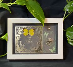 a photo frame with a butterfly on it next to some green leaves and a plant