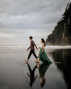 a man and woman walking on the beach with their reflection in the wet sand as they hold hands