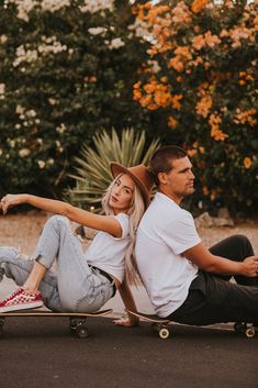 a man and woman sitting on skateboards in the middle of a road with flowers behind them