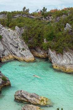 a person swimming in the blue water near some large rocks and trees with green leaves on them