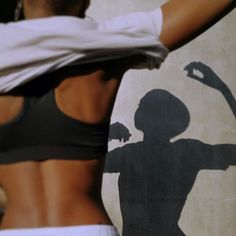 a woman in a sports bra top holding her arms out to the side, while standing next to a wall with a shadow of a man on it