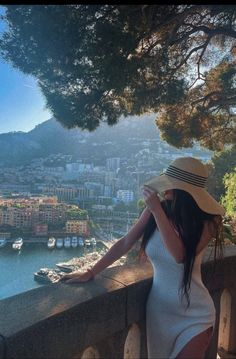 a woman in a white dress and hat leaning on a wall looking at the water