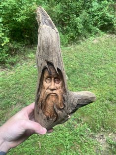 a person holding up a piece of wood with a face on it and trees in the background