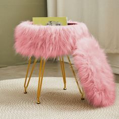 a pink furry stool with gold legs and a card holder on the top, in front of a window