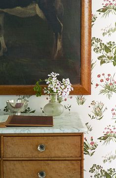 a painting hangs on the wall next to a dresser with flowers in vases and an open book