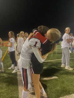 two female football players hugging each other on the sidelines at night with fans in the background