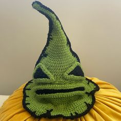 a crocheted hat with a green face on top of a yellow pillow in front of a gray wall