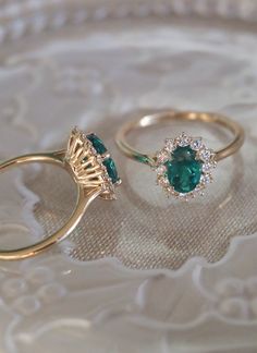 two gold rings with green and white stones on a lace doily, one has a diamond halo