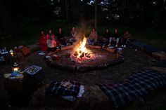 a group of people sitting around a fire pit in the woods at night with blankets on it