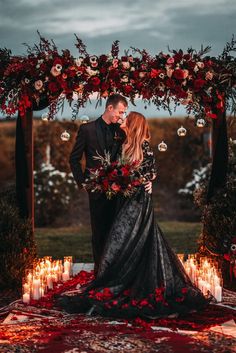 a man and woman standing under an arch with candles on it, surrounded by red flowers