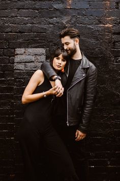 a man and woman standing next to each other in front of a black brick wall